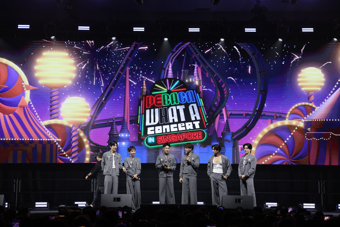 A concert scene featuring six performers on stage, dressed in matching outfits, with a colorful backdrop displaying 'WHAT A CONCERT IN SINGAPORE' against a festive amusement park theme.