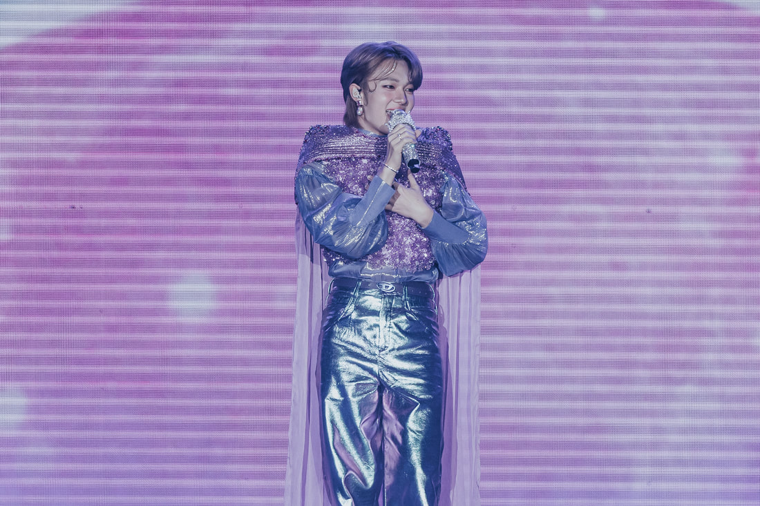A performer singing on stage, wearing a sparkly purple outfit with metallic silver pants and a sheer cape, against a bright pink background.