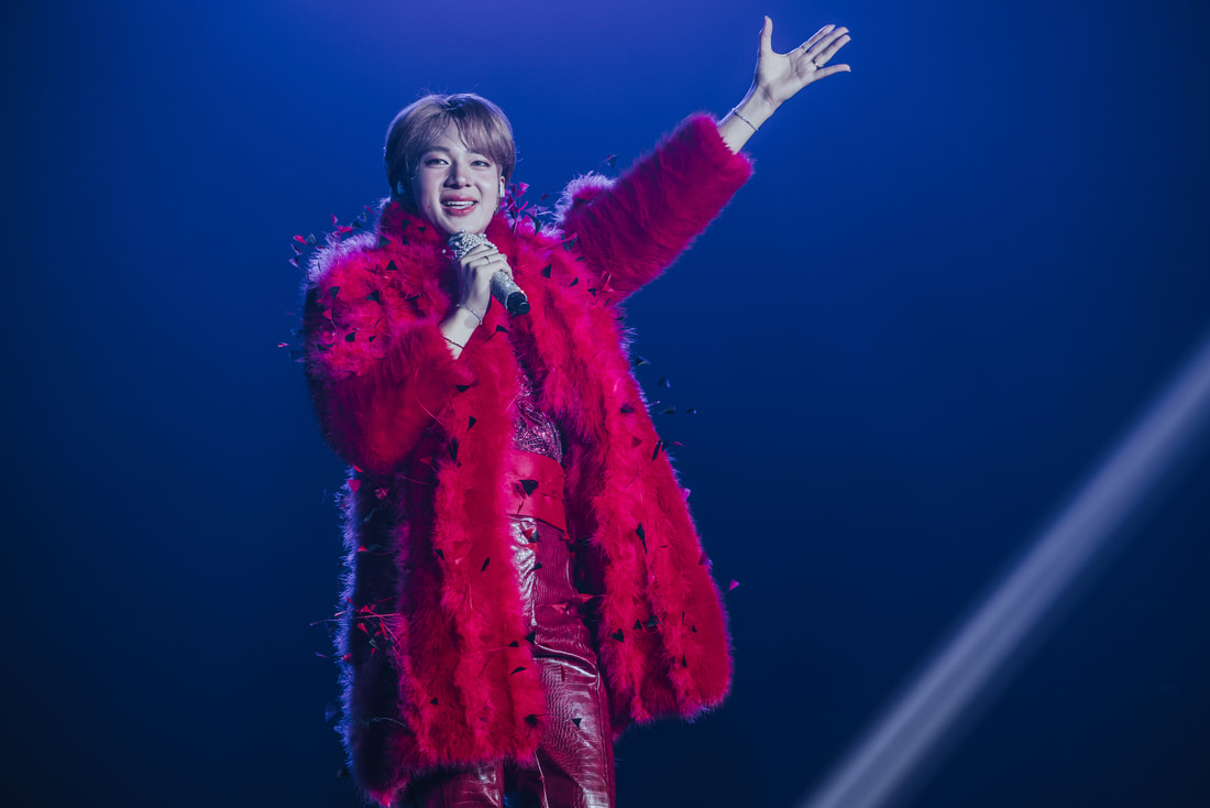 A performer on stage wearing a vibrant red feather coat, holding a microphone and gesturing with one hand in a spotlight.