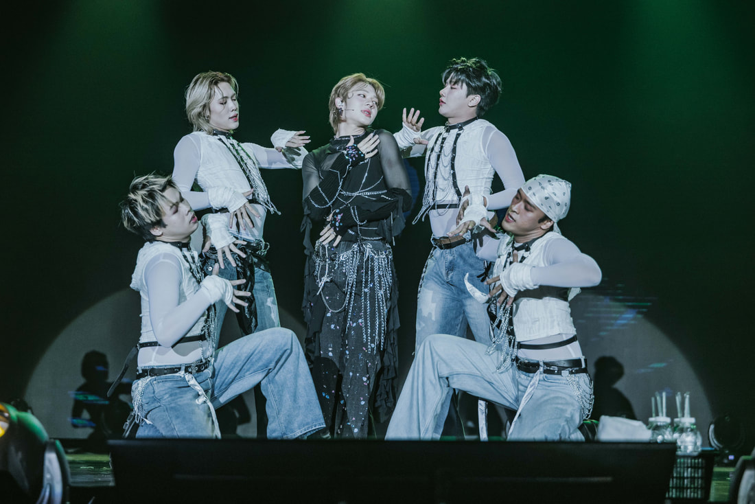 A dynamic performance by five dancers on stage, showcasing elaborate choreography with dramatic poses and stylish outfits, illuminated by stage lighting.