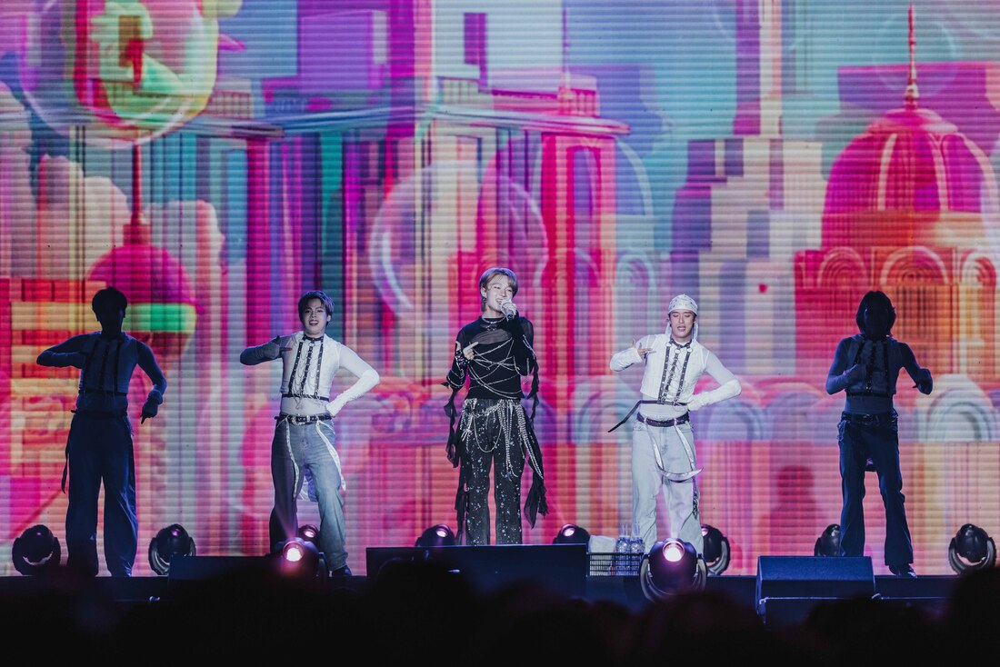 A singer performs on stage surrounded by four dancers, with a colorful digital backdrop depicting abstract buildings and cityscapes.