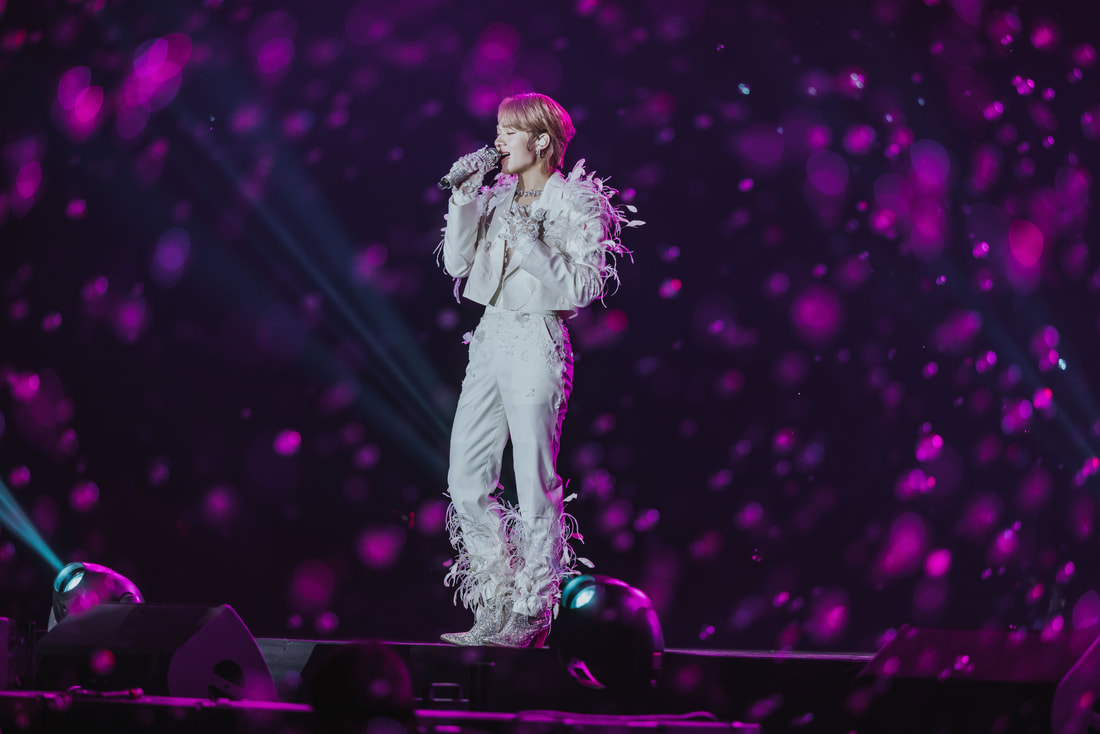 A performer singing on stage while wearing a stylish white outfit with feather details and sparkling boots, surrounded by a colorful light display.