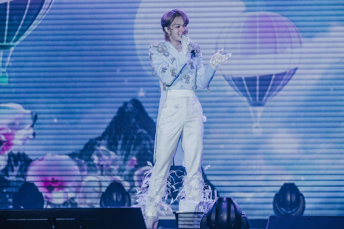 A performer on stage wearing a stylish white outfit adorned with embellishments and feathers, singing into a microphone with a vibrant backdrop featuring hot air balloons.