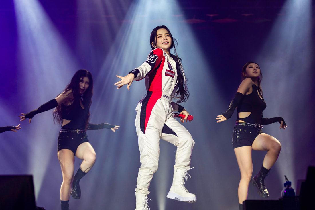 A performer in a racing jumpsuit dances on stage, holding a microphone, surrounded by two backup dancers in stylish outfits, with dramatic lighting in the background.