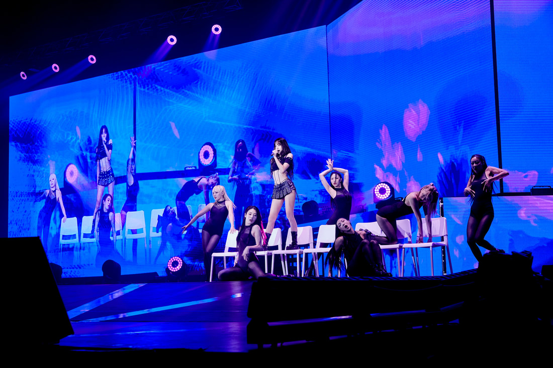 A performer on stage singing passionately with a group of dancers in black outfits and high heels, set against a vibrant blue and pink background with lights. The dancers are posing and interacting around the performer, adding energy to the performance.