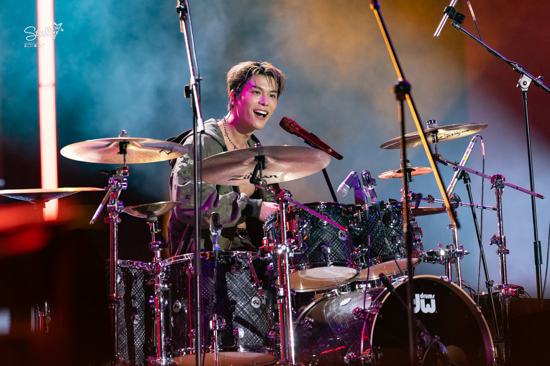A drummer performing on stage, smiling while playing the drums, surrounded by colorful stage lights.