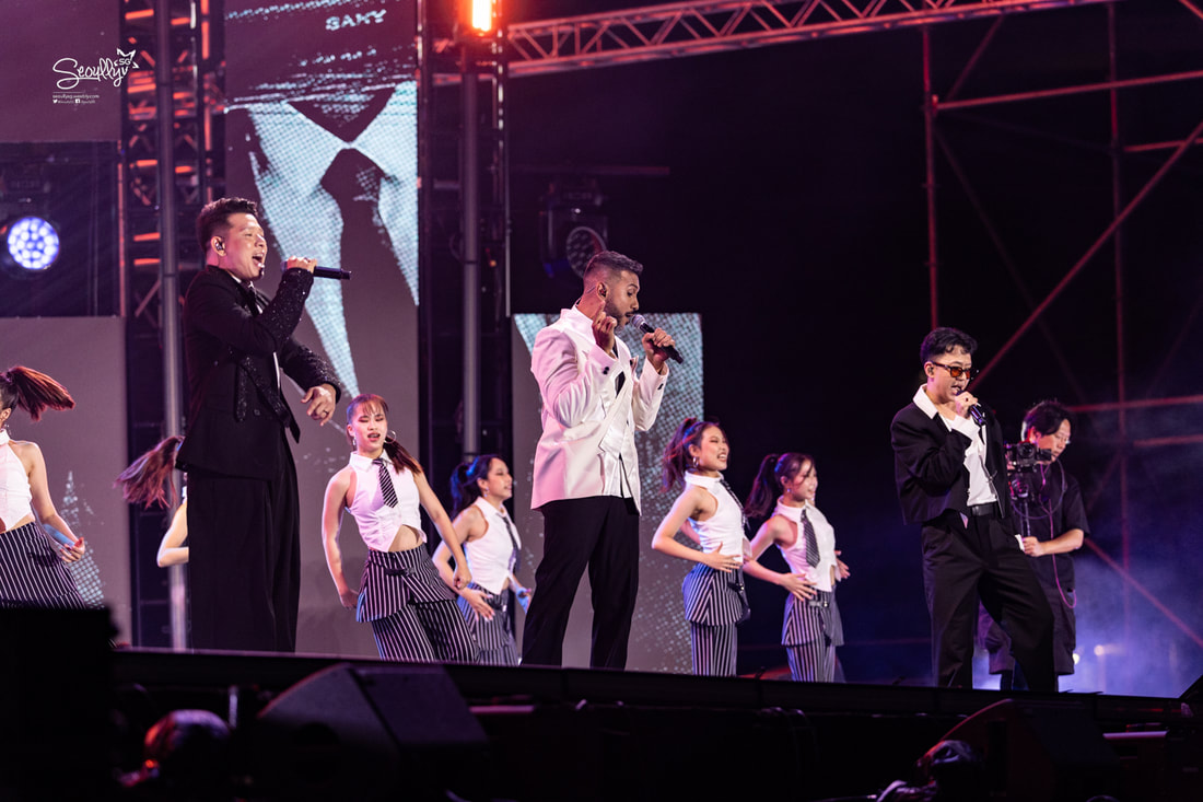 Three male performers singing on stage with dancers in the background during a live concert.