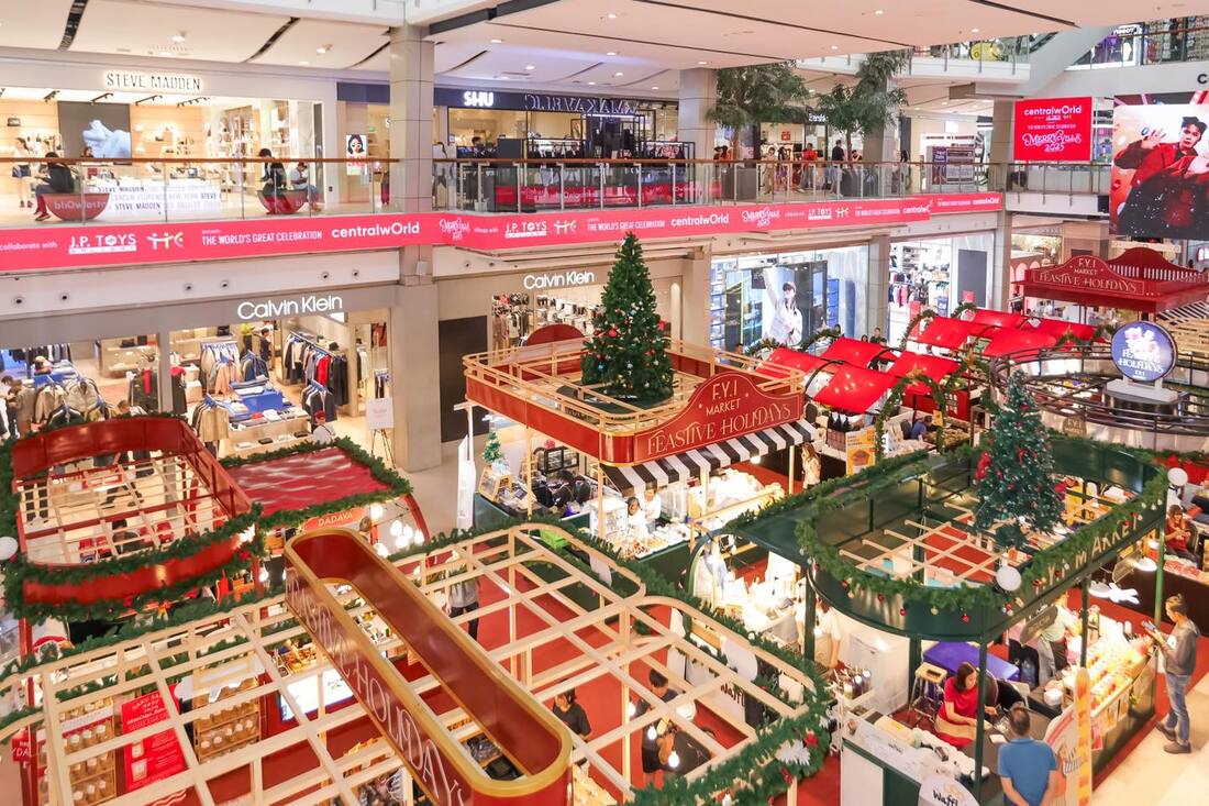 A bustling shopping mall during the festive season, featuring a vibrant holiday market with decorated stalls, Christmas trees, and shoppers exploring various stores like Calvin Klein and Steve Madden.