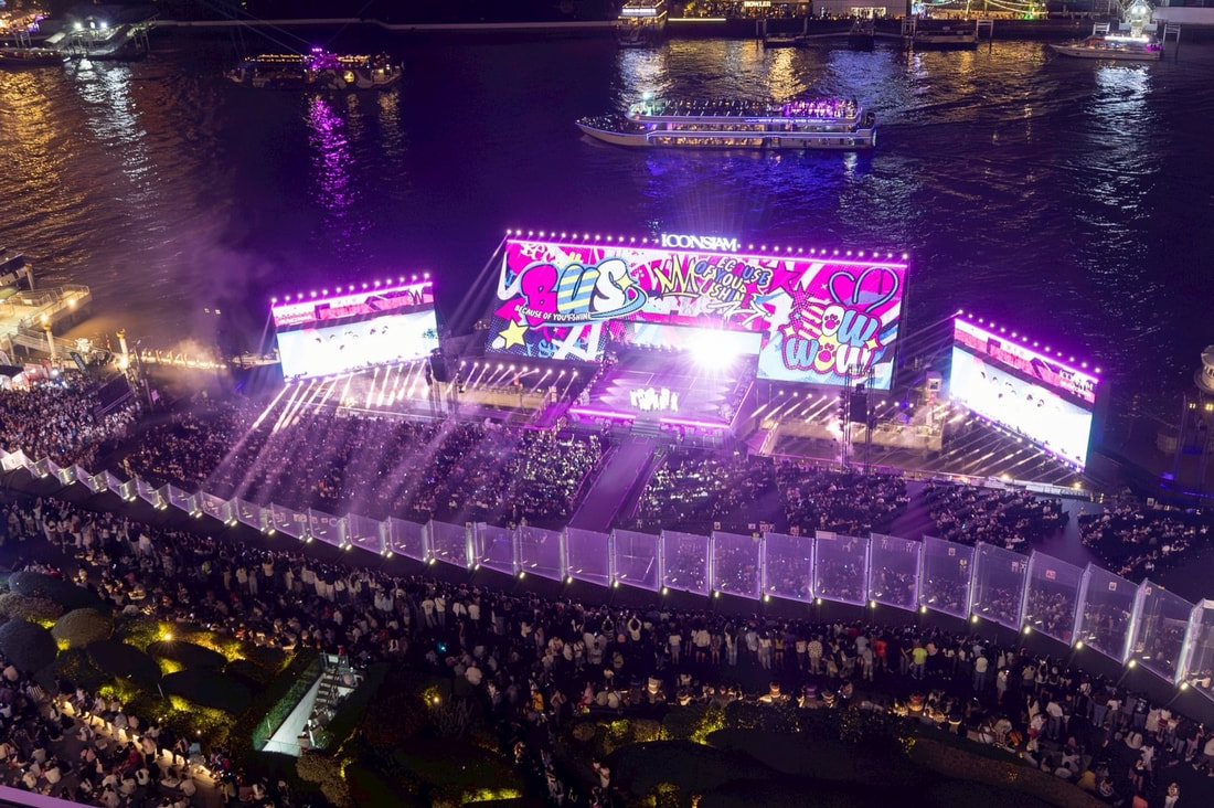 Aerial view of a vibrant outdoor concert with a large stage illuminated by colorful lights, surrounded by a cheering crowd and a river in the background.