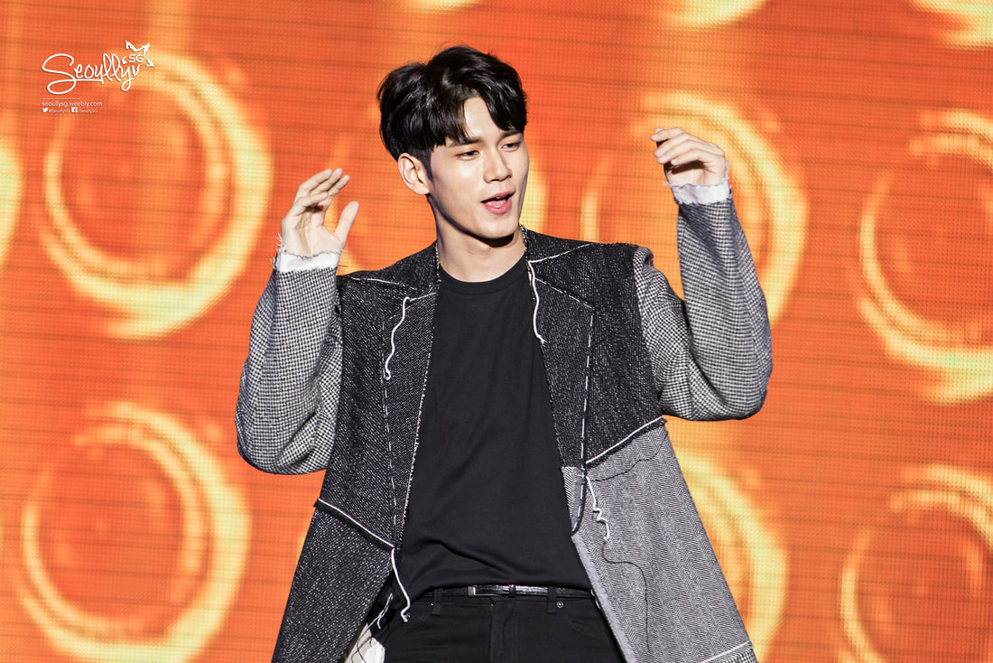 A male performer on stage, wearing a stylish grey and black jacket with a black shirt underneath, enthusiastically gesturing with his hands. The background features vibrant orange circular patterns.