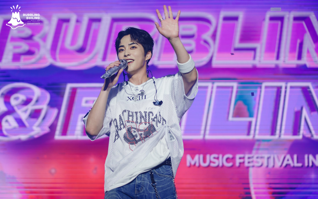 A performer on stage with a microphone, waving to the audience, wearing a white mesh shirt and denim jeans, with colorful lighting in the background and the words 'Bubbling & Boiling Music Festival' visible.
