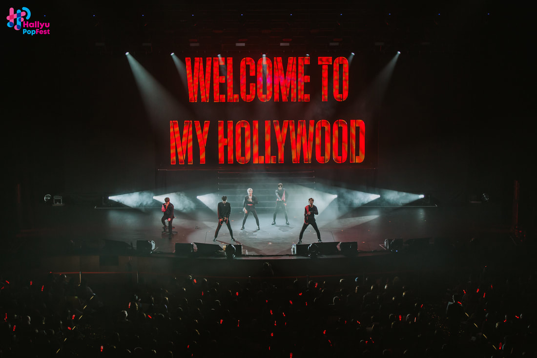 A live concert scene featuring five performers on stage with dramatic lighting. A large screen displays the text 'WELCOME TO MY HOLLYWOOD' in bright red. The audience is visible in the foreground, holding up red light sticks.
