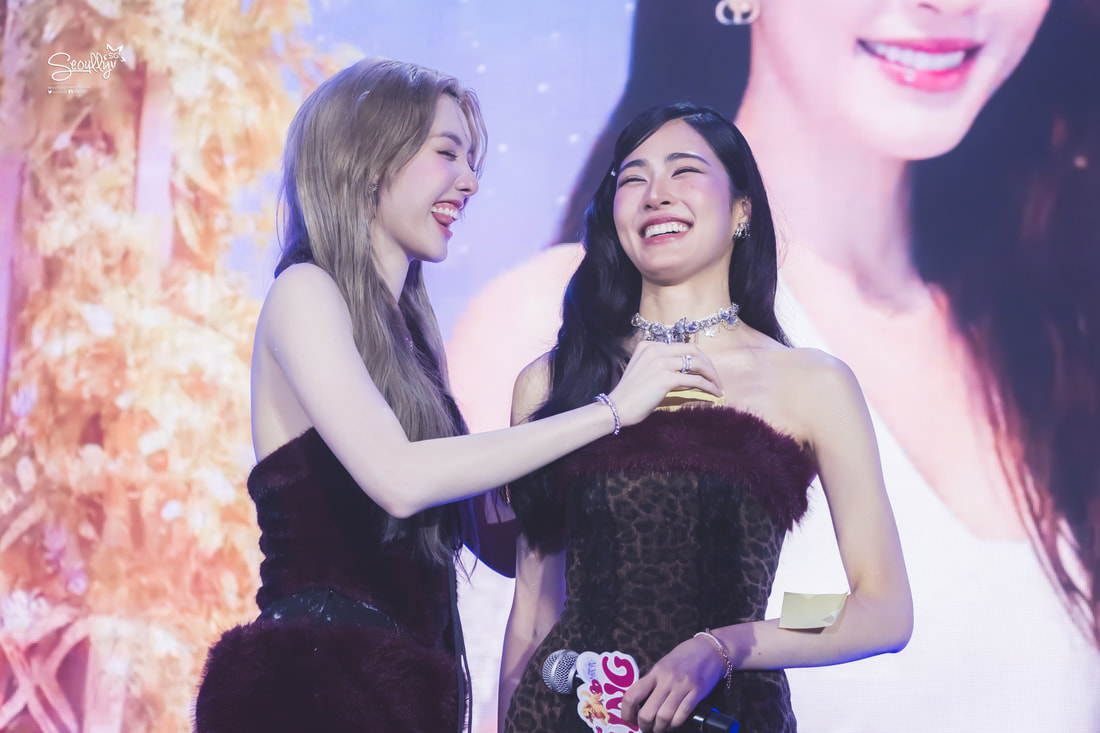 Two women sharing a joyful moment on stage, one playfully touching the other's outfit while both are smiling.