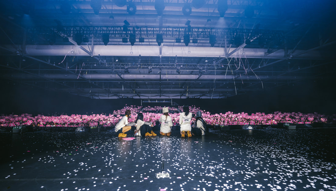 A group of five individuals sitting on a stage, facing an audience filled with pink flower props. The stage is decorated with scattered confetti, and the venue is dimly lit.