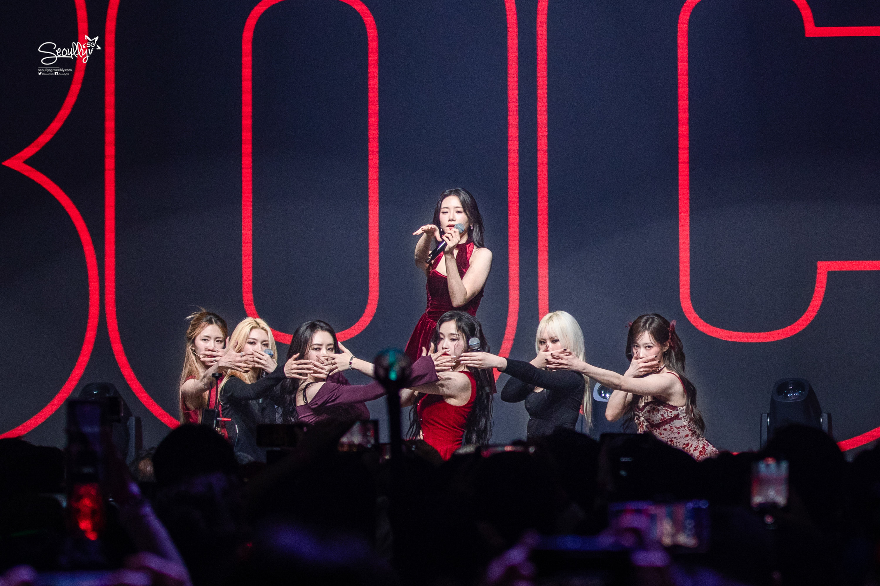 A group of female performers on stage during a concert, with one singer in a red dress in the foreground and others in various outfits, all striking dramatic poses. Bright red lighting and a large number 3010 in the background enhance the performance atmosphere.