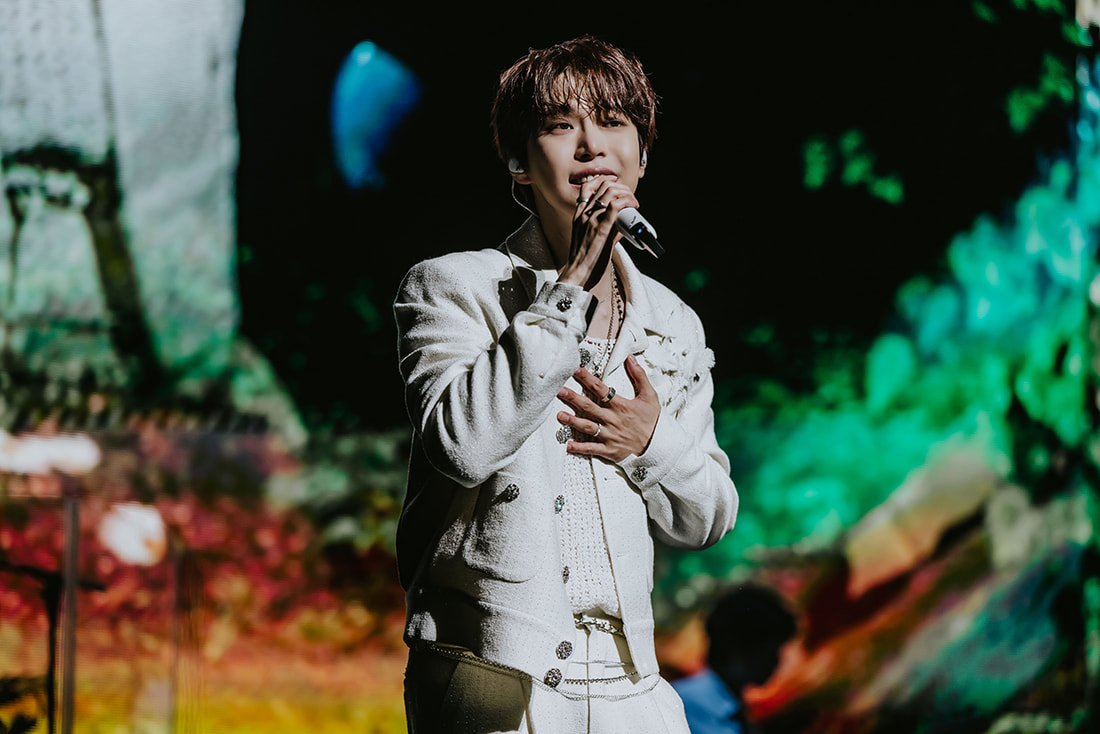 A male singer performing on stage, wearing a stylish white outfit and holding a microphone, with colorful visual effects in the background.