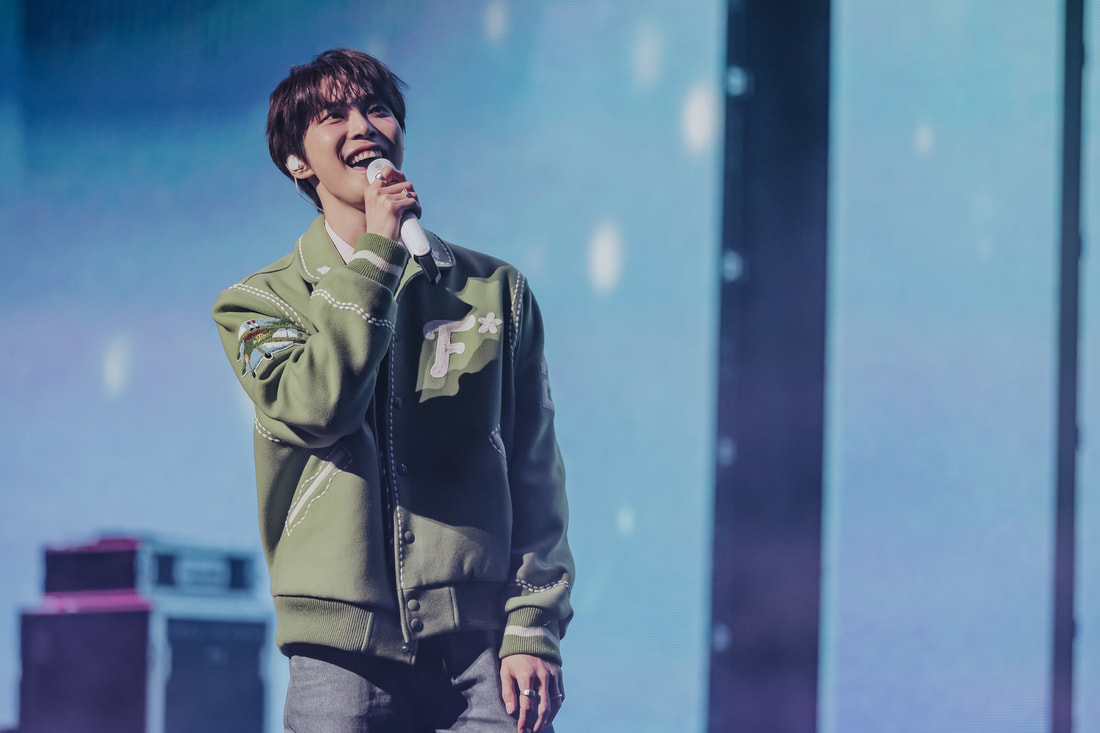A young male singer smiling widely while holding a microphone on stage, wearing a green jacket with decorative patches against a colorful background.
