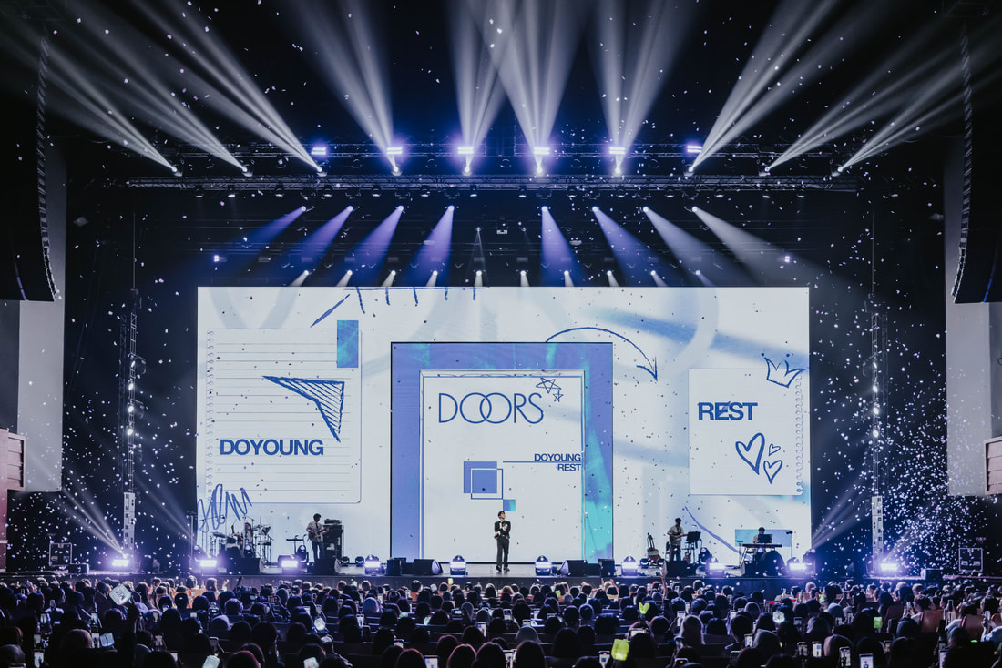 A live performance on stage with a large screen displaying the words 'DOORS', 'DOYOUNG', and 'REST'. The stage is illuminated with bright lights and confetti falling, while musicians are seen performing, and an audience filled with fans is enjoying the show.