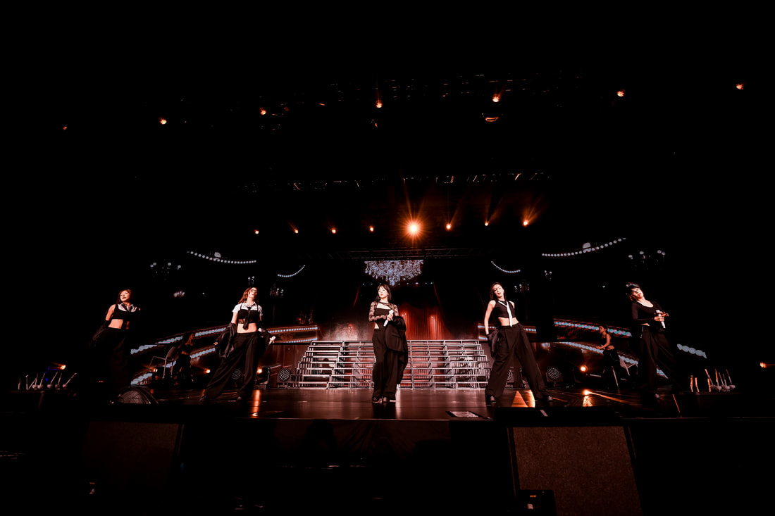 A group of four performers on stage during a live concert, dressed in stylish black outfits with dramatic lighting and a large backdrop.