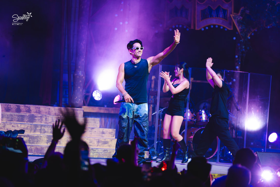 A performer in a sleeveless black shirt and sunglasses waves to the audience during a live concert, accompanied by two dancers in stylish outfits under colorful stage lights.
