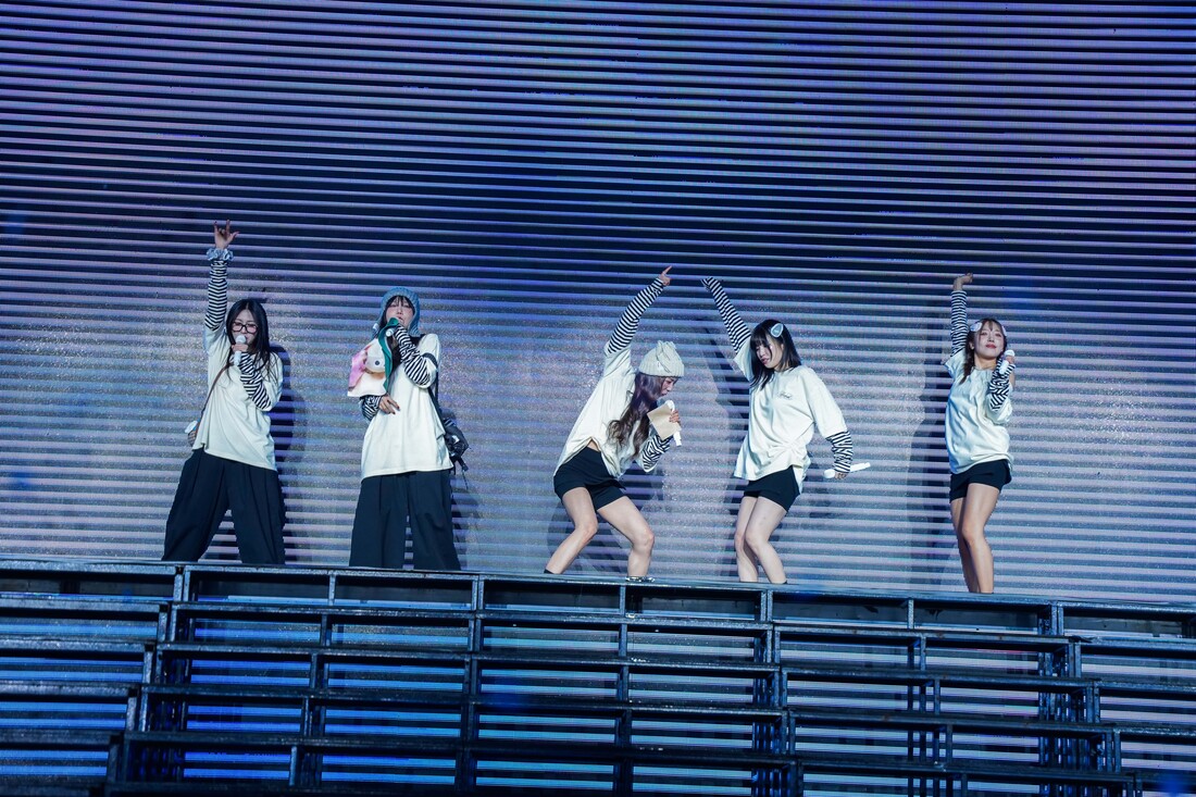A group of five performers dancing on stage, wearing matching outfits with striped sleeves, some holding microphones, under colorful stage lights.