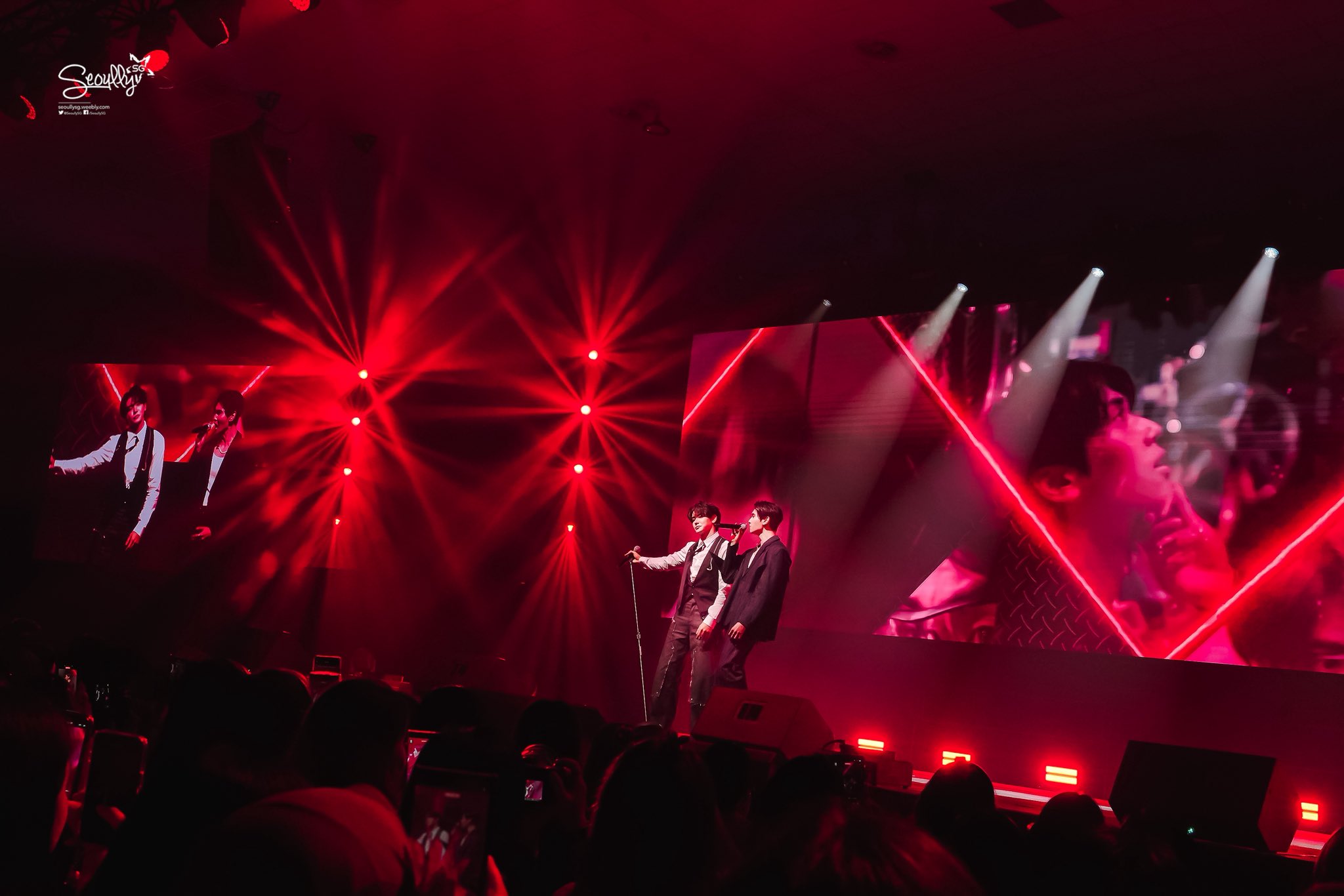 Two performers on stage during a concert, illuminated by red stage lights, with a large screen displaying visuals in the background.