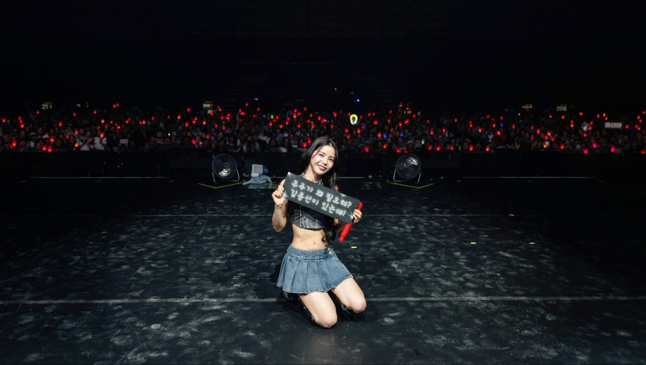 A female performer kneels on stage holding a sign, with a crowd of fans holding red light sticks in the background.