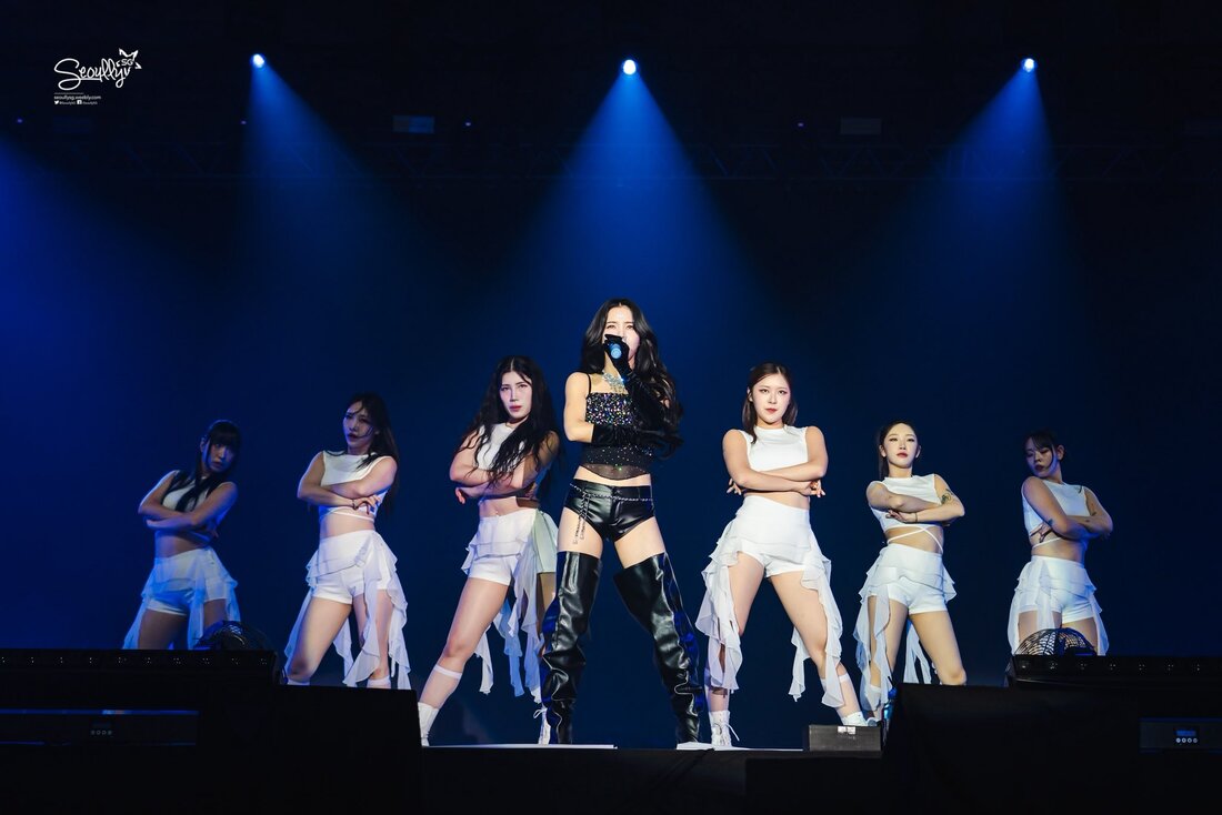 A female singer stands in front of a group of backup dancers on stage, all wearing stylish outfits, with dramatic lighting enhancing the performance atmosphere.