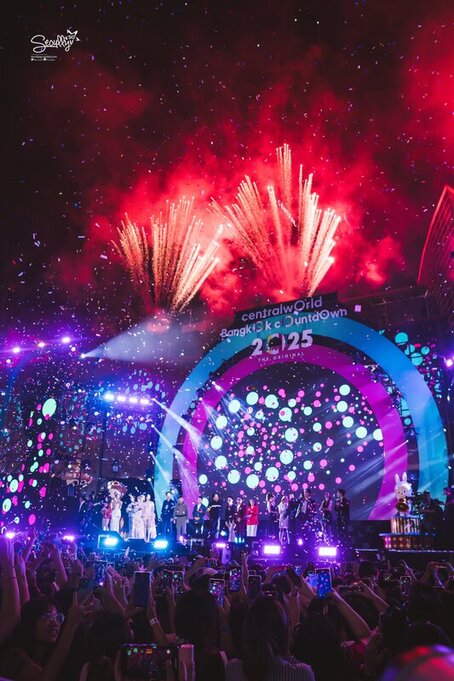 A vibrant New Year's countdown celebration at CentralWorld, Bangkok, featuring colorful fireworks, a lively stage with performers, and a large crowd holding smartphones, with colorful lights and decorations in the background.