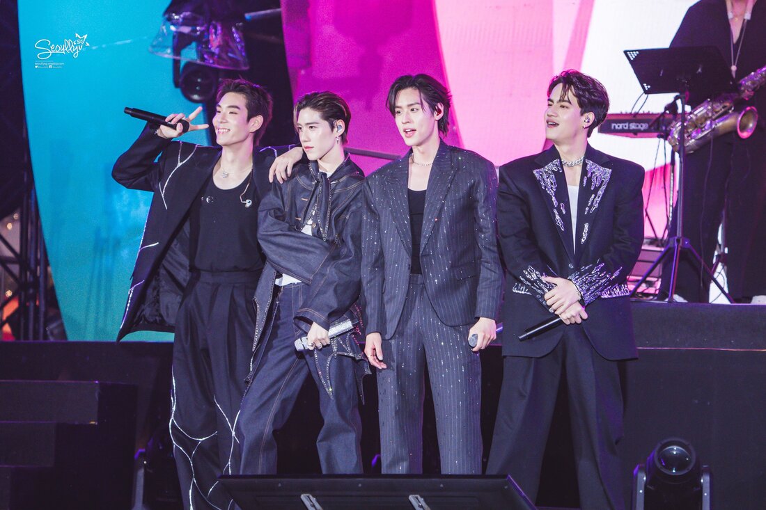 Four male performers on stage, smiling and posing together, with colorful lights in the background.