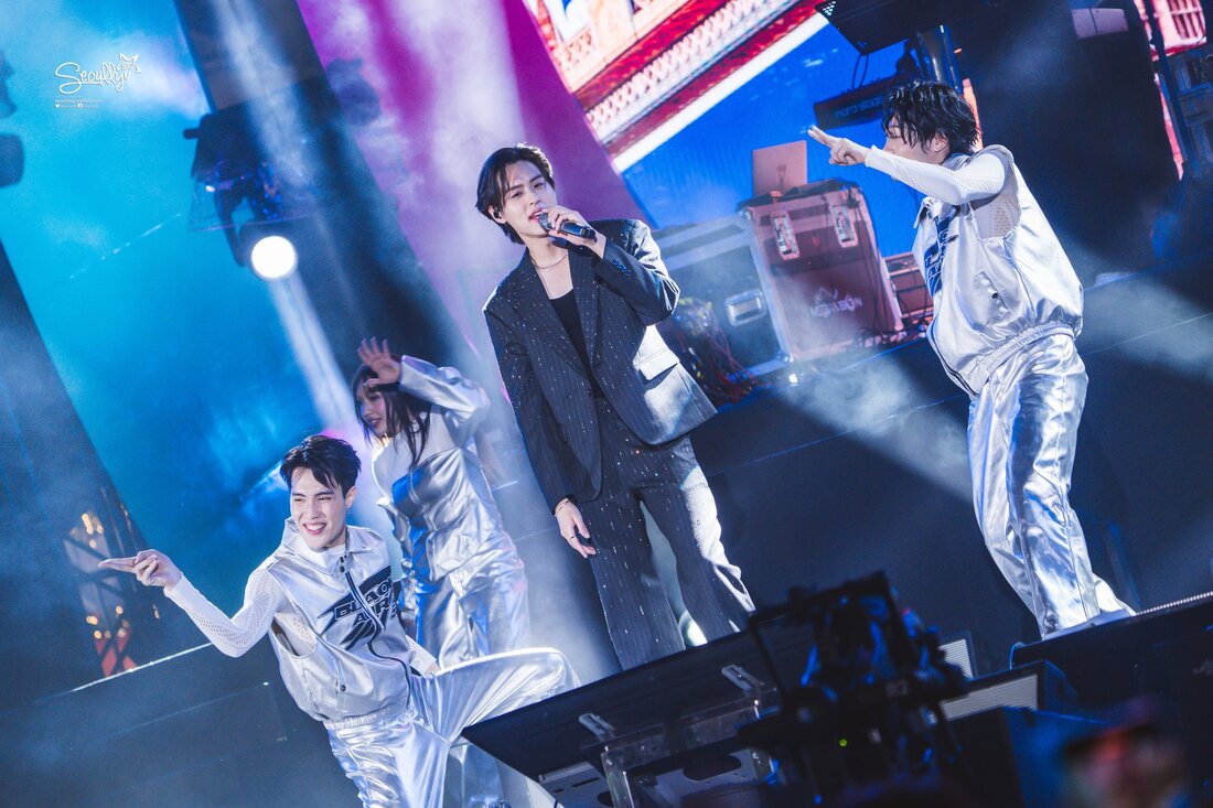 A performer singing on stage surrounded by dancers in shiny outfits, with colorful lights and fog in the background.