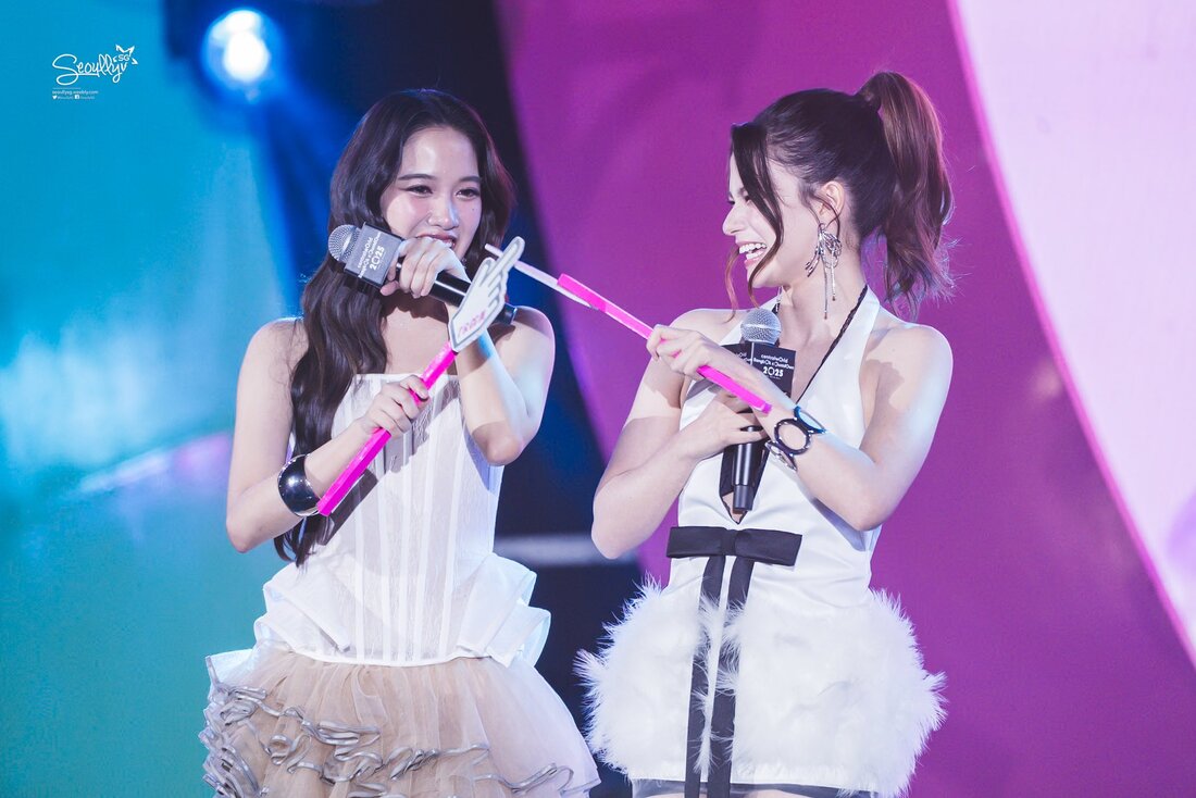 Two women in stylish outfits joyfully interacting on stage, holding microphones and playful props, with vibrant lighting in the background.