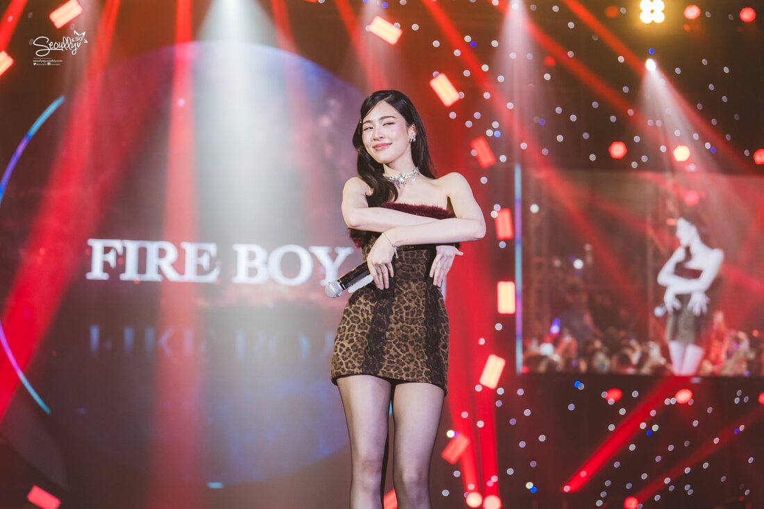 A performer in a leopard print dress and microphone poses on stage, with a colorful background featuring lights and a graphic that reads 'FIRE BOY'.