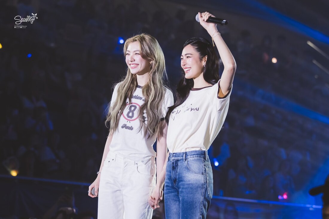 Two female performers smiling and holding hands on stage, with a microphone in one hand and an enthusiastic audience in the background.