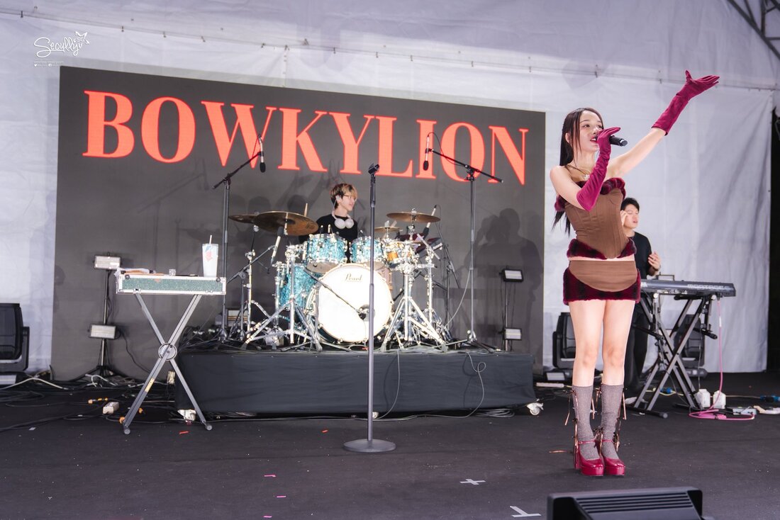 A live music performance featuring a female singer with long hair on stage, dressed in a stylish outfit, singing into a microphone with her arm raised. Behind her, a drummer plays on a brightly colored drum set, while a keyboard player is also visible in the background. The large backdrop displays the name 'BOWKYLION' in bold letters.