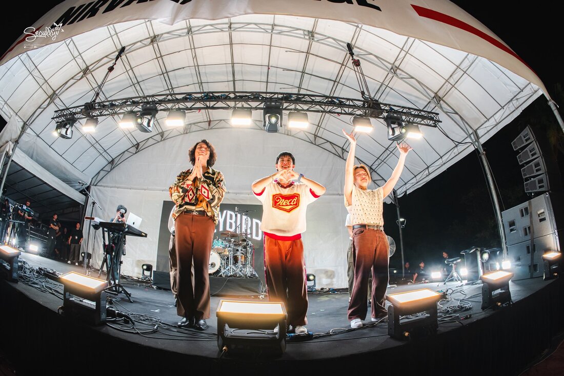 Three performers on stage during a live music event, expressing excitement with gestures. The background features musical instruments and stage lighting.