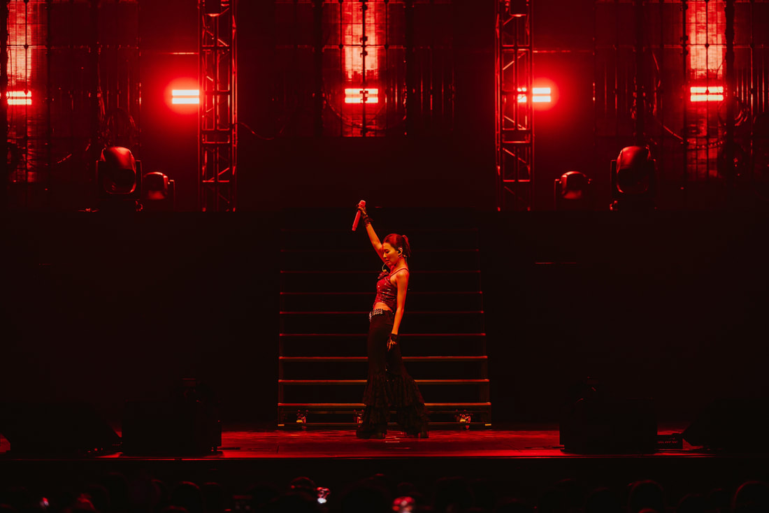 A performer on stage holding a microphone with one arm raised, illuminated by red stage lights, standing in front of a staircase.