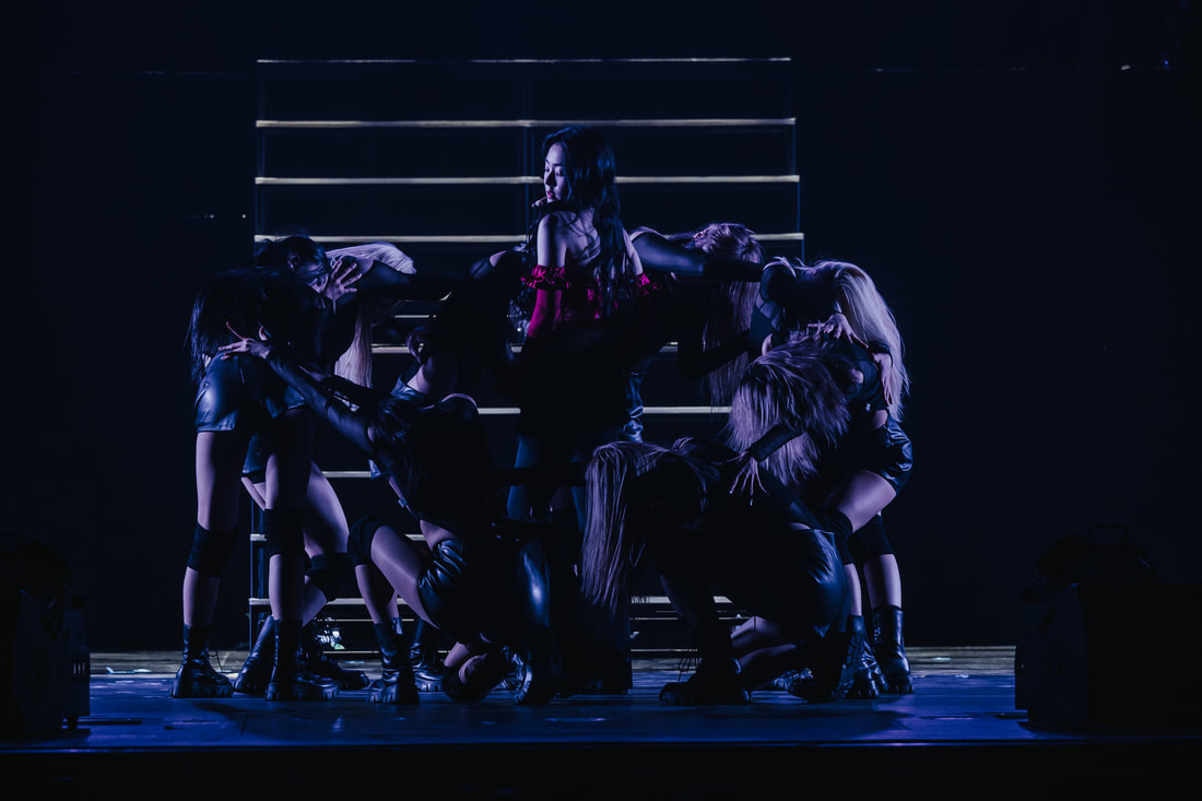 A female performer in a red outfit surrounded by backup dancers in dark clothing, performing on a dimly lit stage with glowing steps in the background.