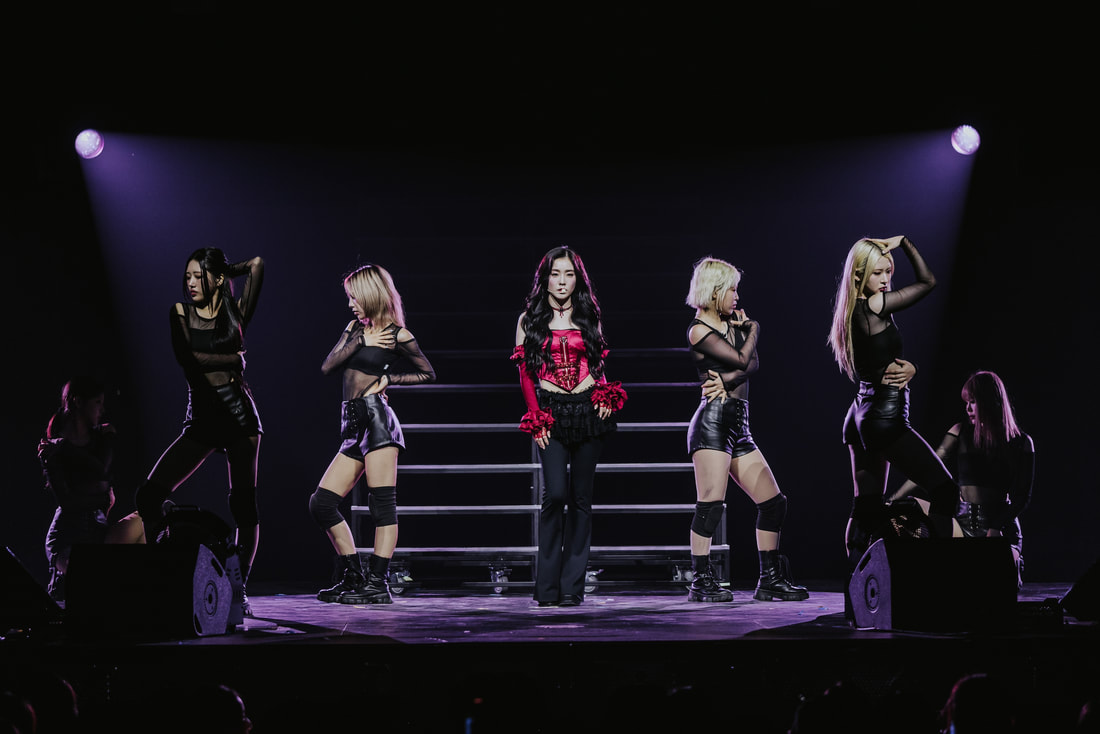 A performer in a red and black outfit stands center stage surrounded by four dancers in black outfits, showcasing a dynamic performance with spotlight effects.