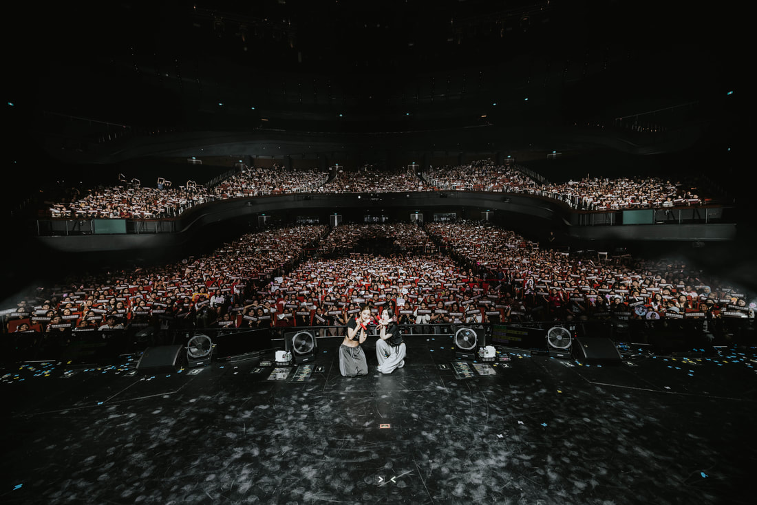 A view from the stage showcasing a large audience holding up banners, with two performers in the foreground, captured during a live concert.