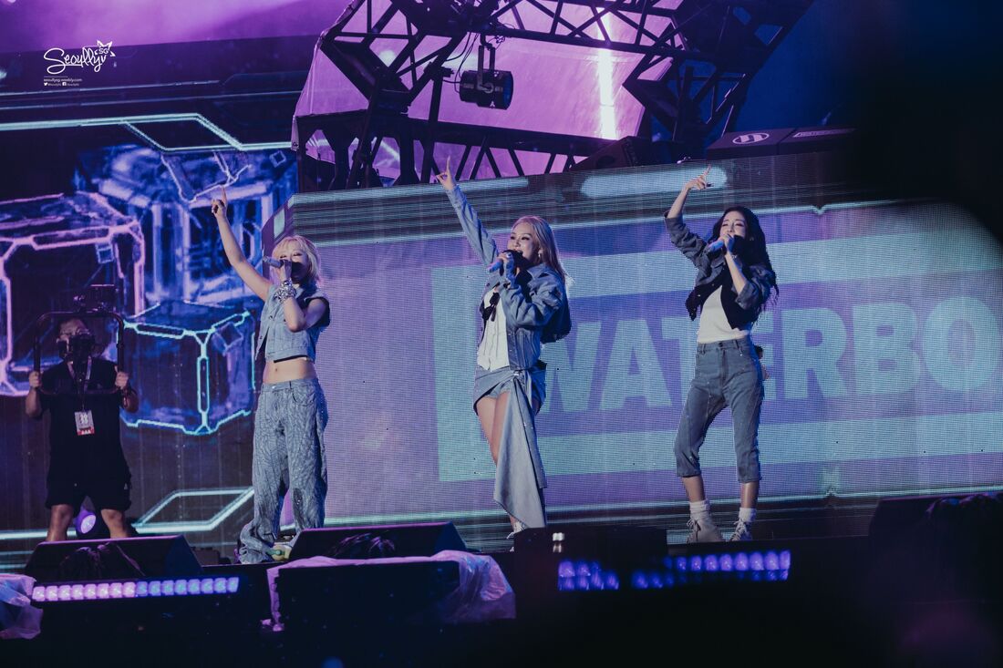Three female performers on stage at a concert, wearing denim outfits and engaging with the audience, with colorful lighting and a large screen in the background.