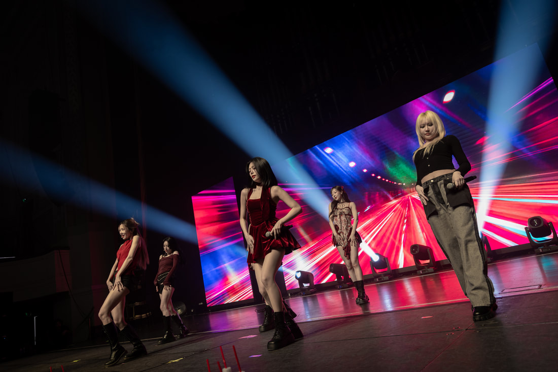Five performers dancing on stage with bright lights and a colorful backdrop during a live performance.