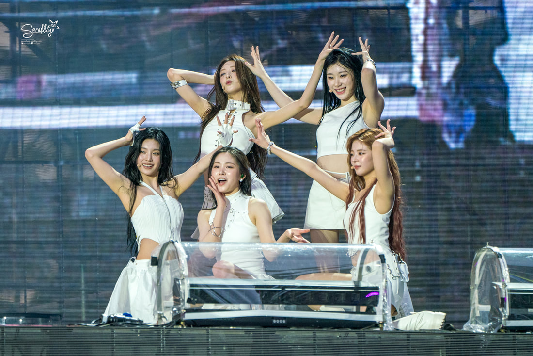 Five female performers dressed in white outfits pose dynamically on stage during a live performance.