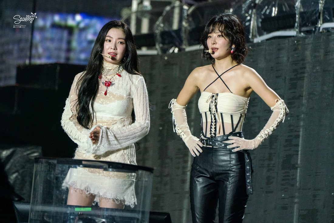 Two female performers on stage during a concert, one in a white lace dress with a red necklace and the other in a black and white outfit with leather pants and gloves.