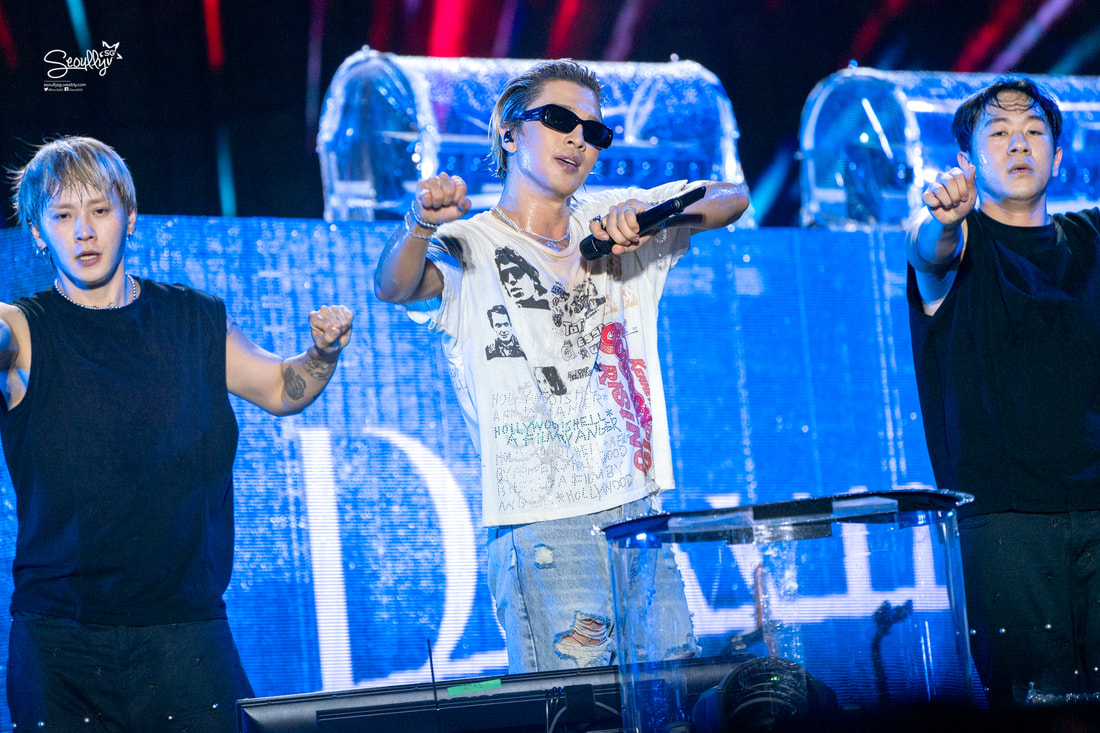 Three male performers on stage during a concert. One is wearing sunglasses and a graphic t-shirt, while the others are in black sleeveless shirts. They appear to be dancing with energetic expressions, with colorful stage lighting in the background.