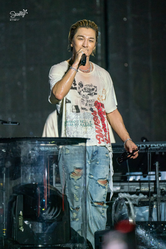 A male singer on stage holding a microphone, wearing a distressed white t-shirt and ripped jeans, with long hair and a confident expression.