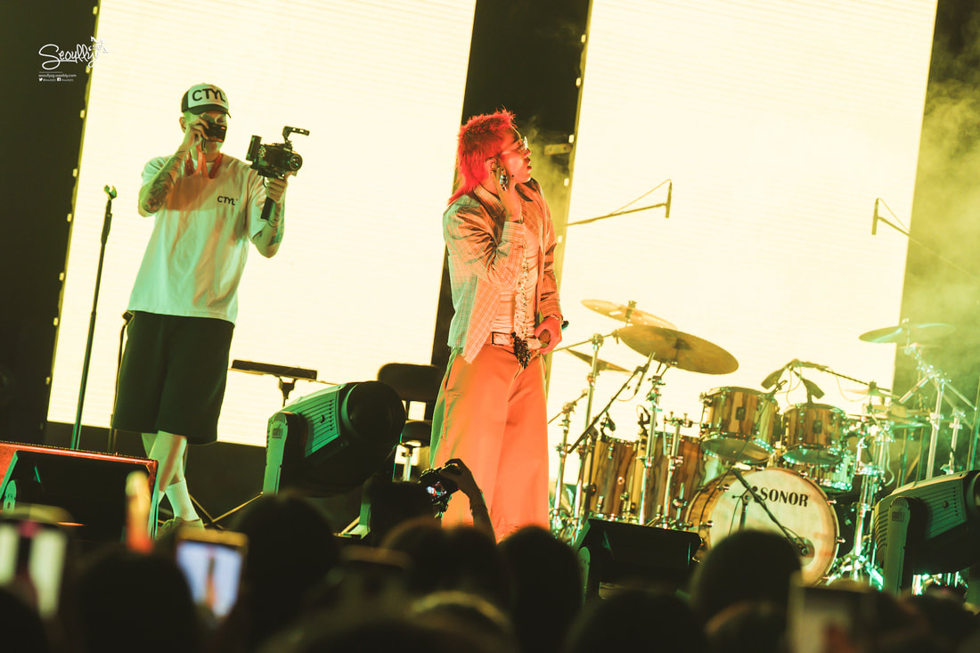 A performer with colorful hair singing on stage, while a cameraman captures the moment in front of an audience, with large screens illuminated in the background.