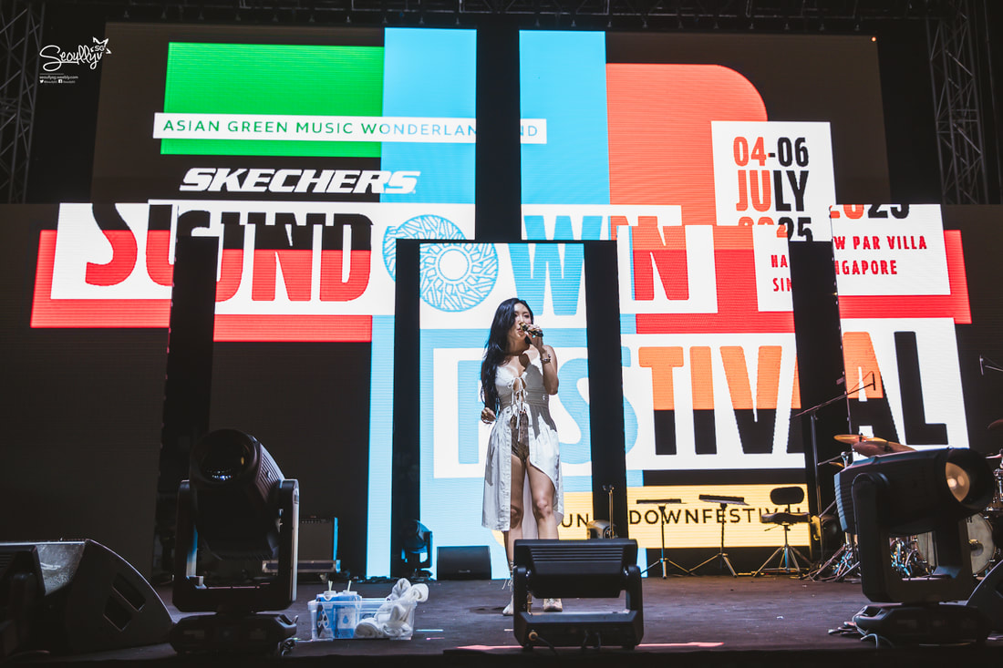 A performer on stage at the Sound Down Festival, with a vibrant backdrop featuring colorful event graphics and announcements about the festival dates and location in Singapore.