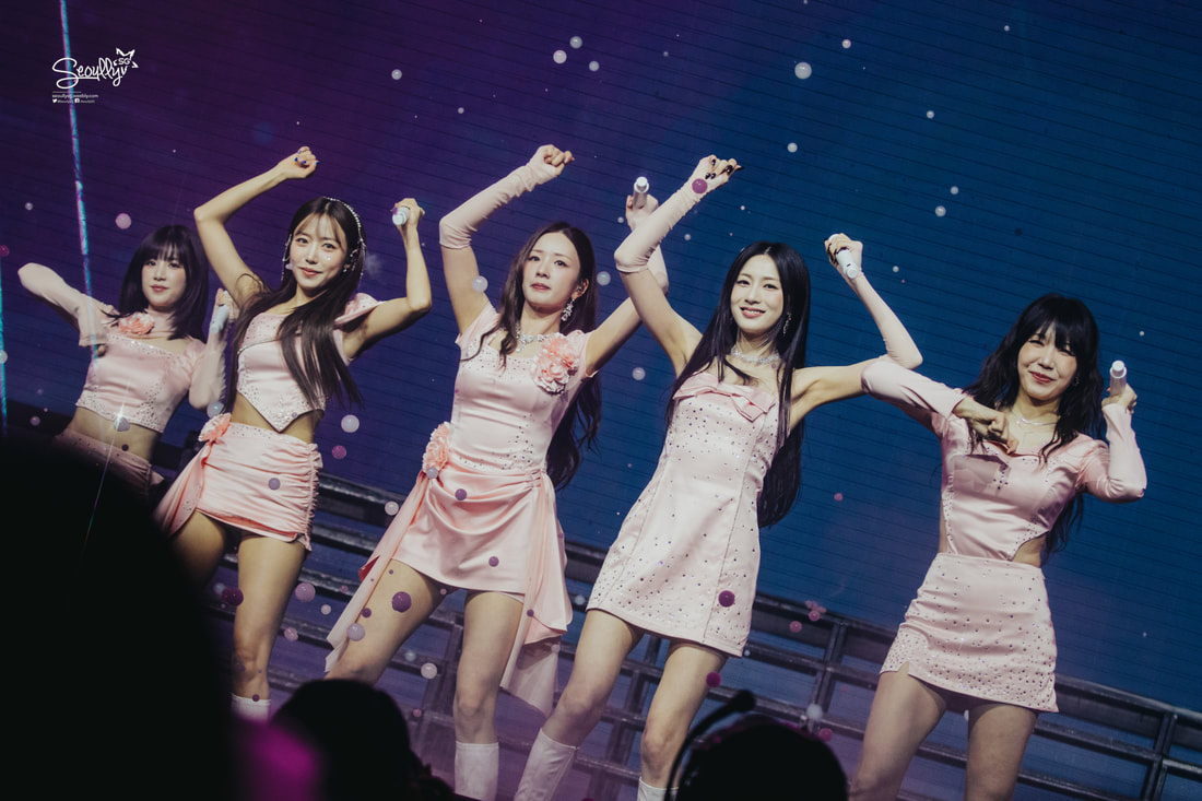 Five female performers in matching pink outfits are dancing on stage, showcasing choreography with joyful expressions. The background is colorful with floating bubbles.