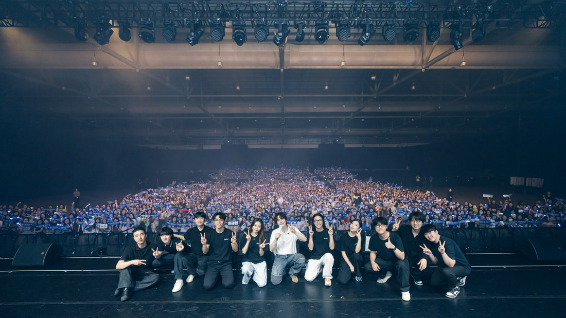 Group of performers posing on stage in front of a cheering crowd at a large event.